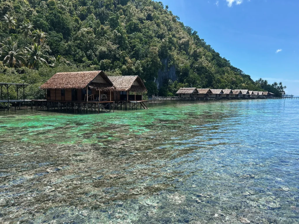 Traditional overwater bungalows with thatched roofs beside a lush tropical island, surrounded by clear turquoise water and coral reef.